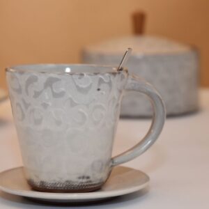 handgetöpferte Keramiktasse mit handgemachten Unterteller aus Keramik in weiß und Vorratsdose aus Keramik im Hintergrund
