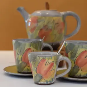 Keramik, Steingut und Steinzeug - Handgetöpferte Keramiktasse mit Löffel und weiteren Keramiktassen und einer Teekanne aus Keramik im Hintergrund.
