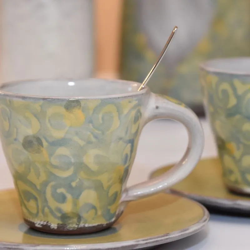 Handgetöpferte Keramik - Handgetöpferte Tasse aus Steinzeug Keramik in grün auf einem Unterteller und weiterem handgefertigtem Keramik Geschirr im Hintergrund.